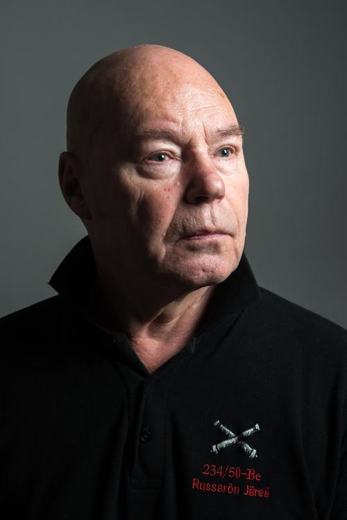 Muotokuva 67-vuotiaasta miehestä, jonka katse on suunnattu oikealle. Mies on kalju ja hänellä on musta pusero, jonka rintamuksessa on ristissä kaksi merikanuunaa. Paidassa on teksti 234/50 Be, Russarön Järeä.